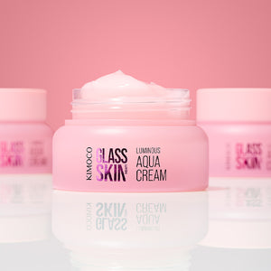 Zdjęcie na różowym tle: po środku otwarty różowy słoiczek "Kimoco Glass Skin Luminous Aqua Cream" z widocznym kremem na wierzchu, z rozmytymi słoiczkami w tle po obu stronach i odbiciem na lustrzanej powierzchni.