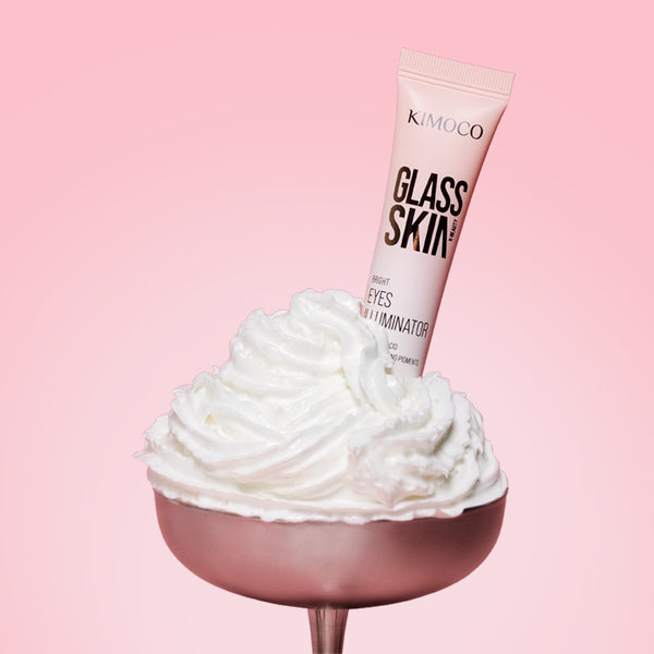 Tuba "Kimoco Glass Skin Bright Eyes Illuminator" wbita w bitą śmietanę ułożoną w metalowym pucharku, na różowym tle.