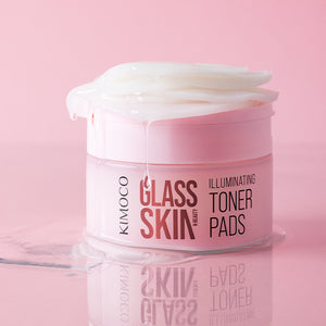 Pojemnik "Kimoco Glass Skin Illuminating Toner Pads" z płatkami wystającymi z wieczka i ściekającym płynem, z odbiciem na lustrzanej powierzchni, na różowym tle.