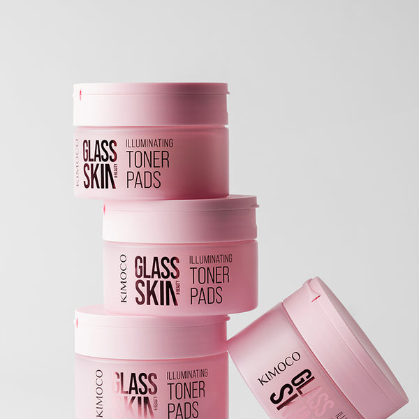 Ustawione w stos pojemniki "Kimoco Glass Skin Illuminating Toner Pads" z jednym pojemnikiem leżącym obok na boku, na jasnoszarym tle.