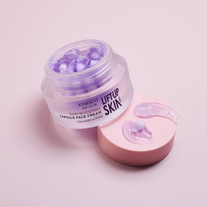 Z lewej otwarty fioletowy słoiczek kosmetyku Kimoco Lift-Up Skin Skin Boosting Capsule Face Cream z widocznymi kapsułkami wewnątrz, z prawej różowa zakrętka z dwiema wyciśniętymi kapsułkami produktu, na jasnoróżowym tle.