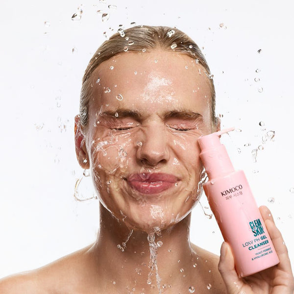 Kobieta z blond włosami i zamkniętymi oczami, z wodą chlapiącą na twarz, trzymająca z prawej strony różową butelkę Kimoco Clean Skin Gentle Cleansing Foam, na białym tle.