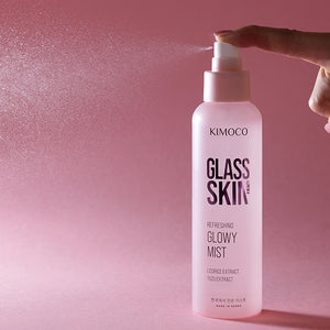 Palce naciskające pompkę różowej butelki kosmetyku Kimoco Glass Skin Refreshing Glowy Mist, z widoczną mgiełką produktu rozpylającą się w lewą stronę, na różowym tle