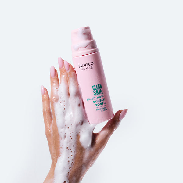 Zdjęcie na jasnoszarym tle: u góry różowa butelka "Kimoco Clean Skin Smoothing Bubble Toner" z pianką na wieczku, u dołu dłoń pokryta białą pianką, unosząca butelkę.