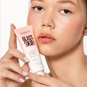 Zdjęcie na białym tle: z lewej dłoń trzymająca tubę "Kimoco Glass Skin Luminous Dew SPF 50+ PA++++" przy twarzy, z prawej twarz kobiety z rumieńcem, patrząca w bok.
