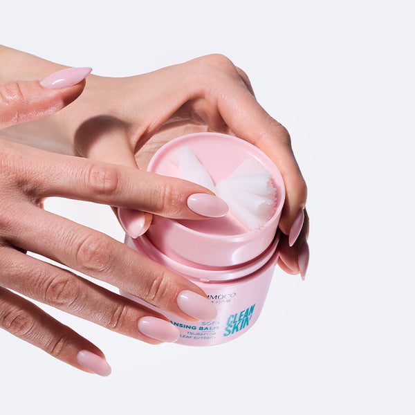 Dwie dłonie z różowym manicure otwierające różowy słoiczek kosmetyku Kimoco Clean Skin Soft Cleansing Balm z białym balsamem w środku, na białym tle.