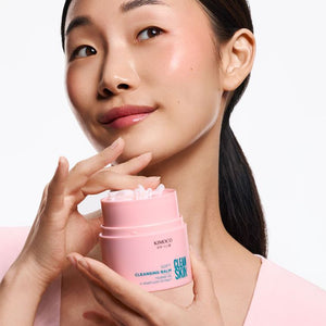 Kobieta o azjatyckich rysach twarzy i ciemnych włosach, trzymająca przed sobą otwarty różowy słoiczek Kimoco Clean Skin Soft Cleansing Balm, dotykająca palcem podbródka, na białym tle.