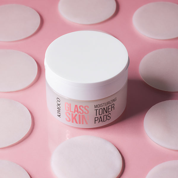 Zamknięty pojemnik "Kimoco Glass Skin Moisturizing Toner Pads" otoczony rozłożonymi okrągłymi płatkami, na różowym tle.