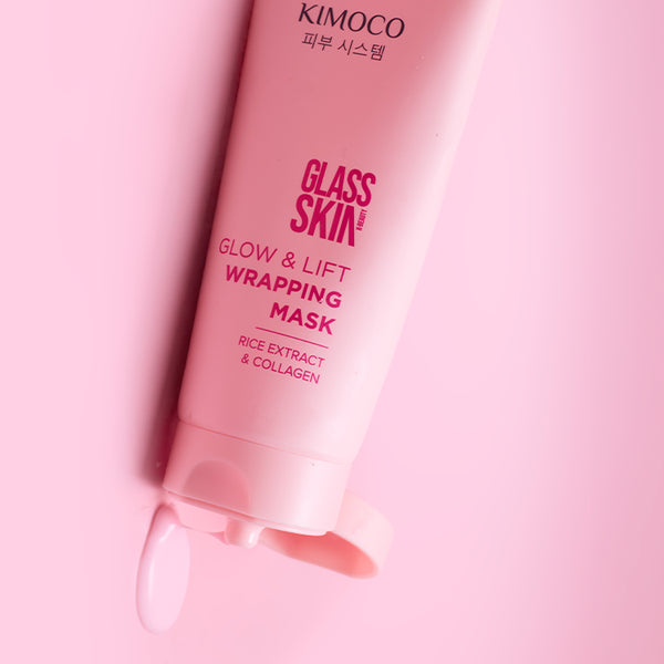 Zbliżenie na różową tubkę kosmetyku Kimoco Glass Skin Glow & Lift Wrapping Mask z ekstraktem ryżowym i kolagenem, z otwartej zakrętki wypływa kropla produktu, na różowym tle