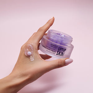 Dłoń trzymająca otwarty fioletowy słoiczek kosmetyku Kimoco Lift-Up Skin Skin Boosting Capsule Face Cream z kolagenem i PDRN, z kapsułką produktu rozsmarowaną na palcu wskazującym, na jasnoróżowym tle.